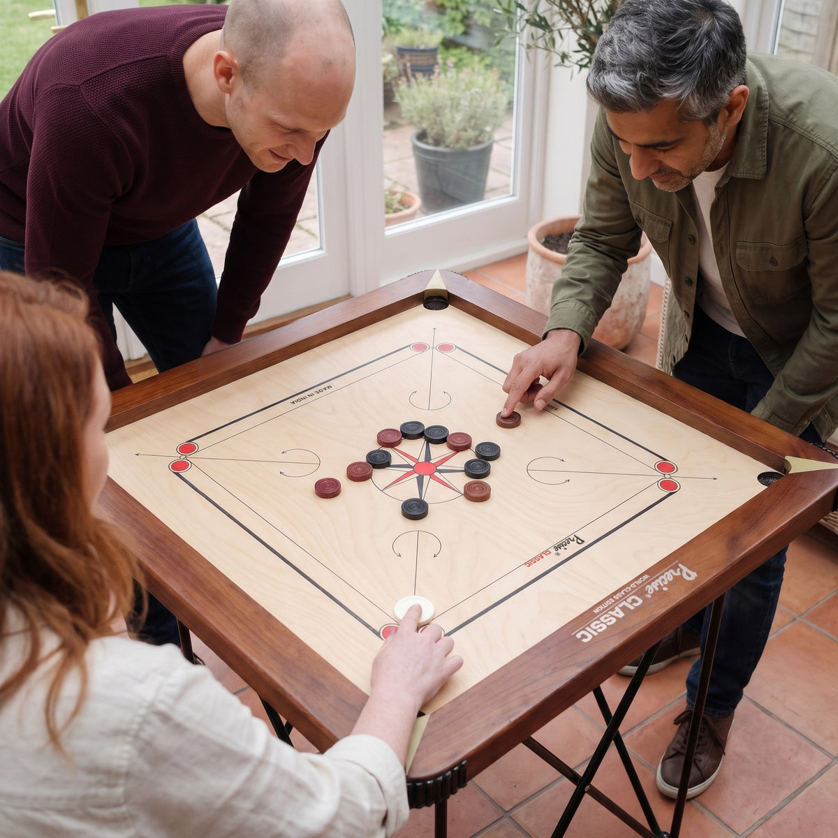Precise Carrom Champion 24mm World-Class Edition van dichtbij: drie vrienden spelen in de tuinkamer, bord met walnootkleurige rand en speelstenen goed zichtbaar